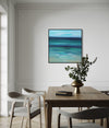 Abstract square landscape artwork depicting a serene seascape in shades of blue and green, displayed on a wall in a modern dining room setting with a wooden table and chairs.