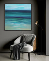 Serene abstract landscape artwork depicting a calming seascape with soft, blurred hues of blue and green. The artwork is framed and displayed on a dark wall, complemented by a cozy armchair in the foreground.