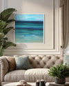 Abstract square artwork depicting a serene seascape in teal and blue hues, framed and displayed on a wall in a cozy, modern living room setting.