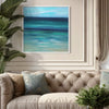 Serene abstract seascape painting in a coastal-inspired living room setting. The large square artwork features a calming, aquamarine-hued landscape against a neutral, tufted sofa and lush, tropical plants, creating a tranquil and inviting atmosphere.